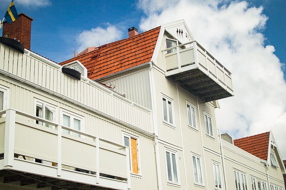 Vit träfasad med balkonger och tegeltak mot blå himmel, en svensk flagga syns vid huset.